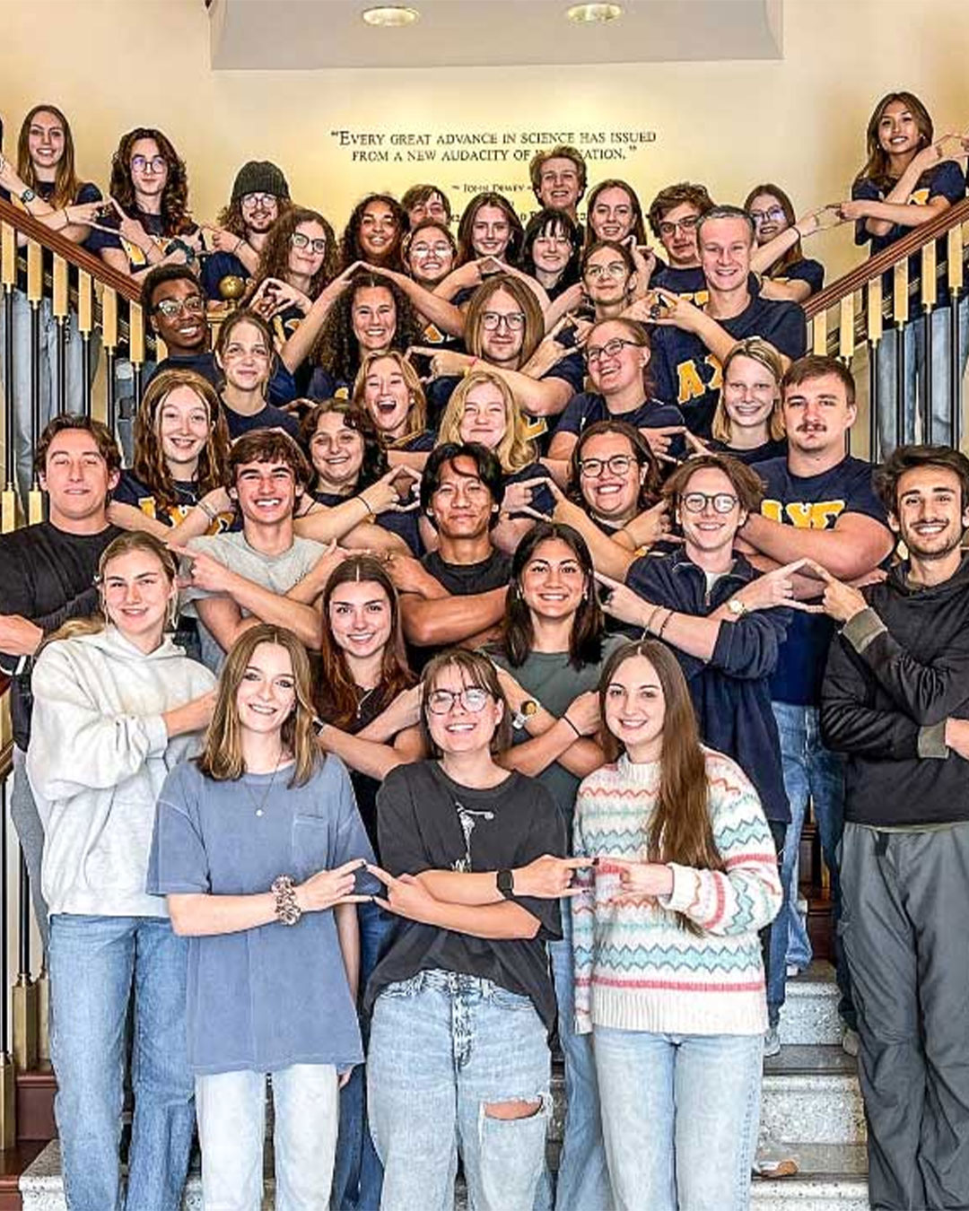 Christopher Newport's chapter of Alpha Chi Sigma poses for a group photo on a grand staircase and flashes their hand sign.
