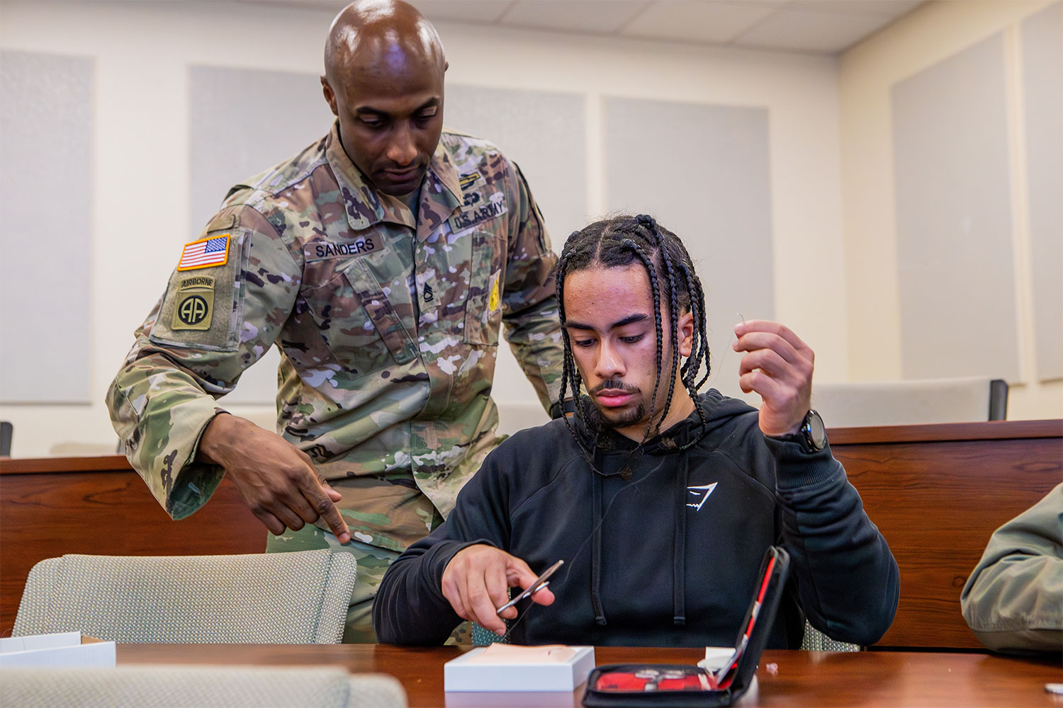 Gemini said An Army soldier guides a premed student practicing suturing techniques on a medical training pad in a classroom.