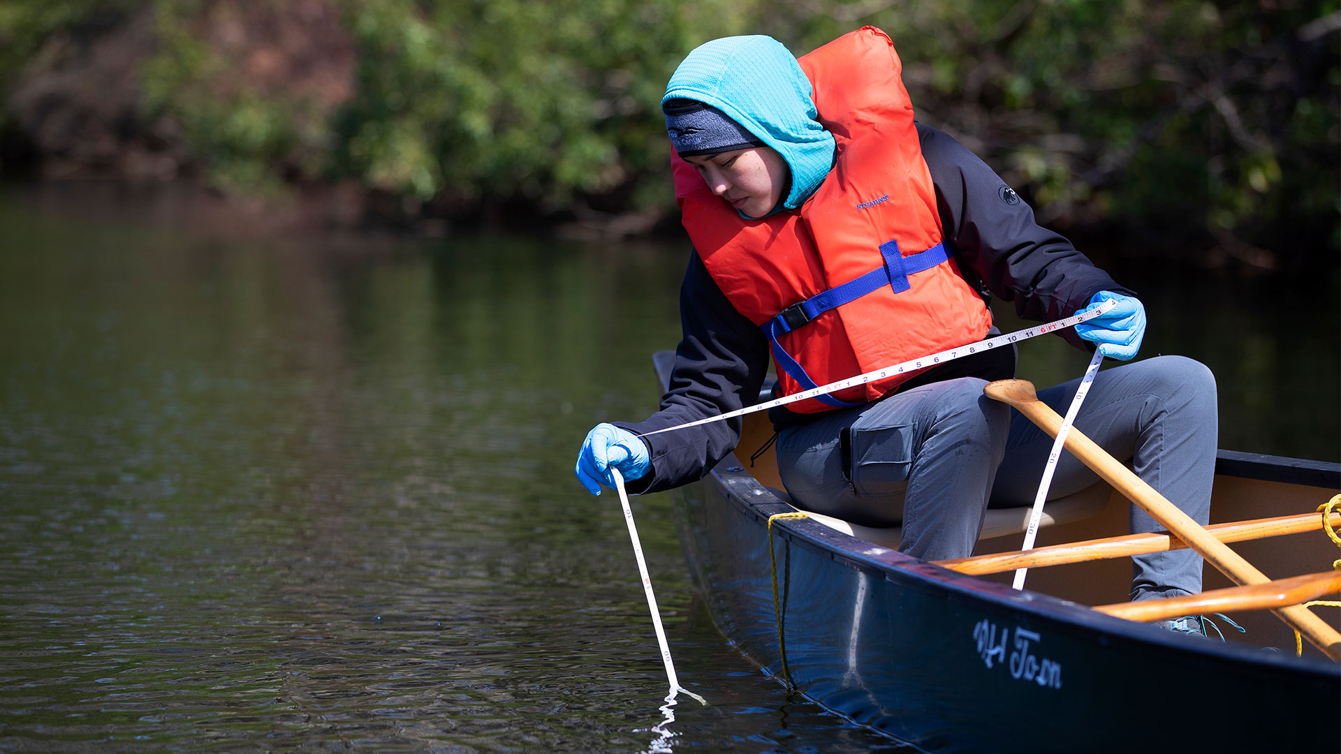 A Christopher Newport ecology student sits in an Old Town canoe and uses a measuring tape to gauge the water depth during fieldwork on a sunny day.