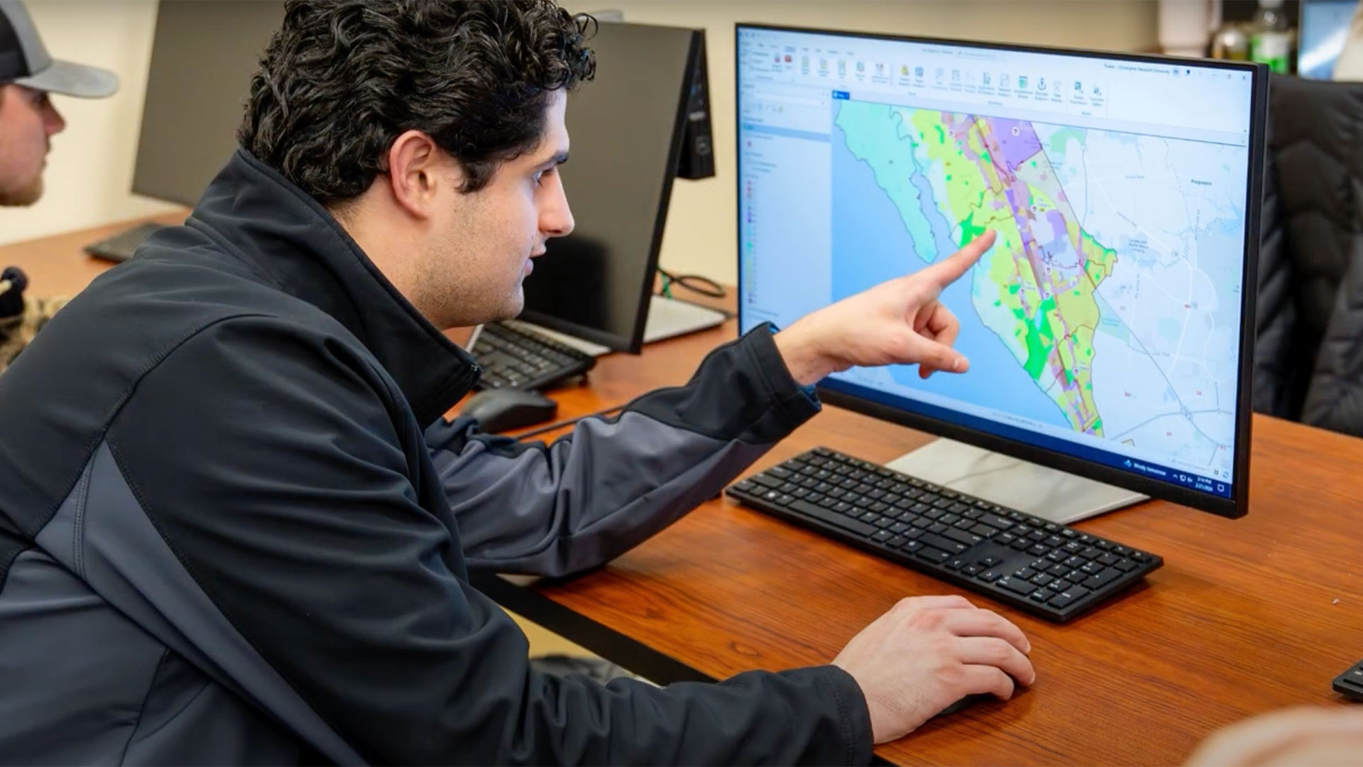 A male student or researcher sits at a wooden desk in front of a computer, pointing at a GIS map displayed on the screen. The map shows different colored regions overlaid on a street grid. He is wearing a dark jacket and is focused on the screen, with a keyboard in front of him. Another person is partially visible to the left.