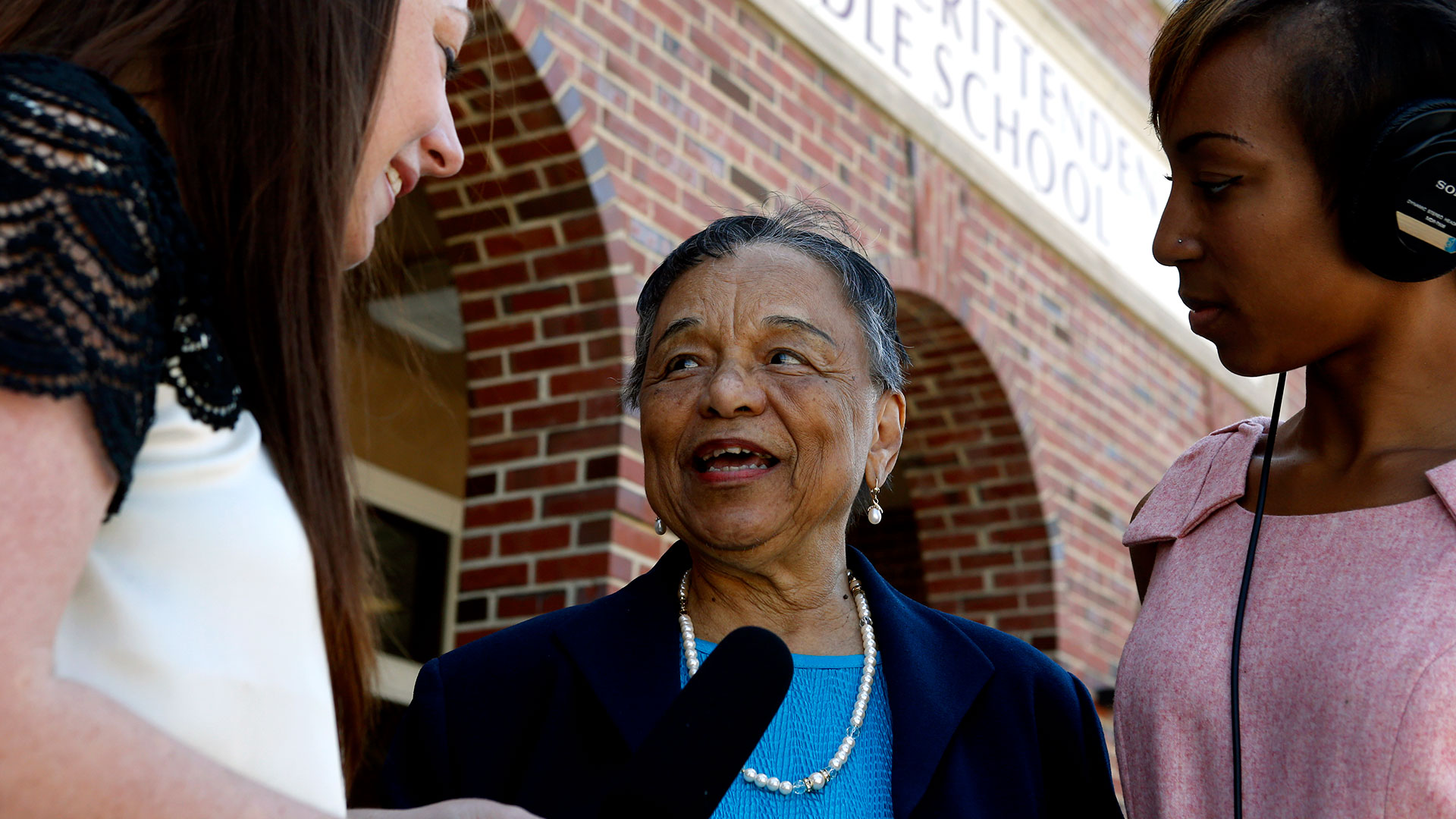 A portrait-style photo of Flora Crittenden, an older woman, standing between two younger women, smiling and looking off-camera. She is wearing a dark jacket over a blue top and a pearl necklace, and is holding a microphone near her chin. The scene takes place outside in front of a brick building with CRITTENDEN MIDDLE SCHOOL visible above her head. One younger woman on the right is wearing large headphones.