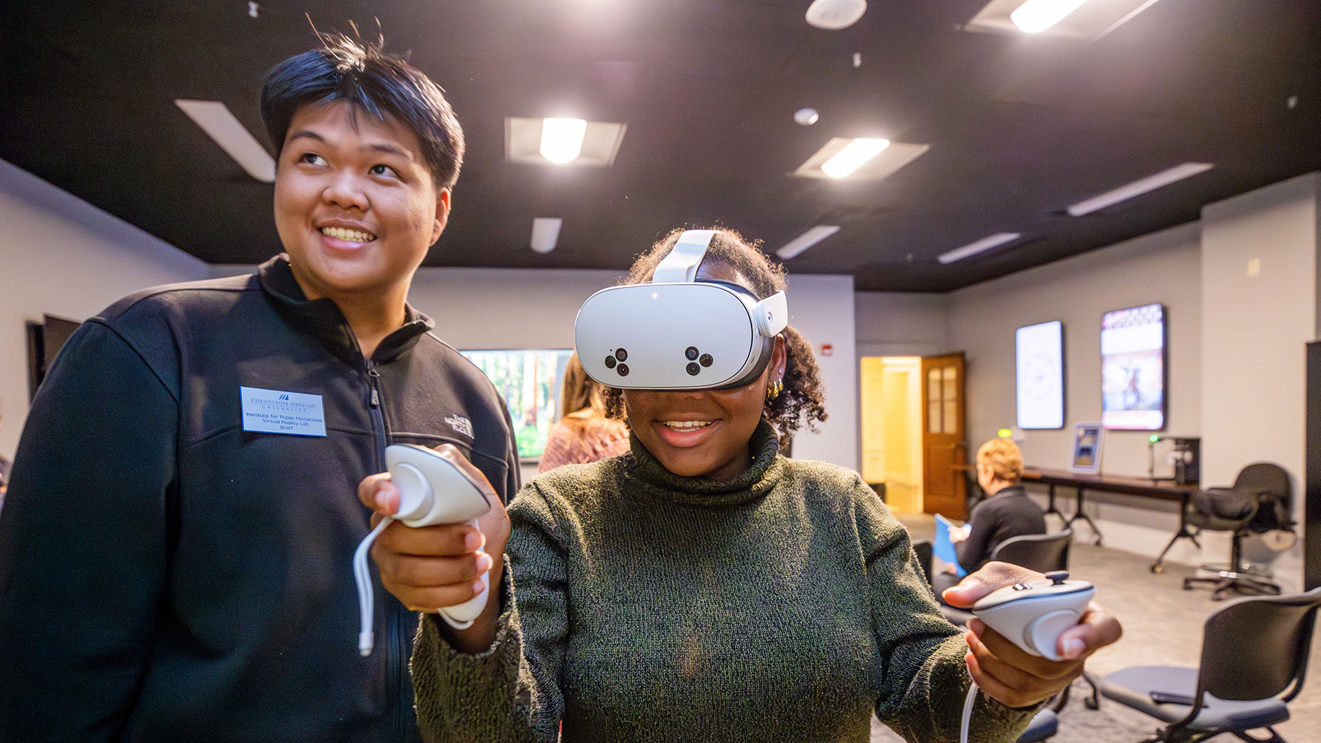 Two young adults are in a room with a black ceiling and bright lights. A Black woman is smiling and wearing a white virtual reality (VR) headset and holding two VR controllers, actively engaged in a VR experience. A man with a nametag is standing next to her, looking on and smiling.