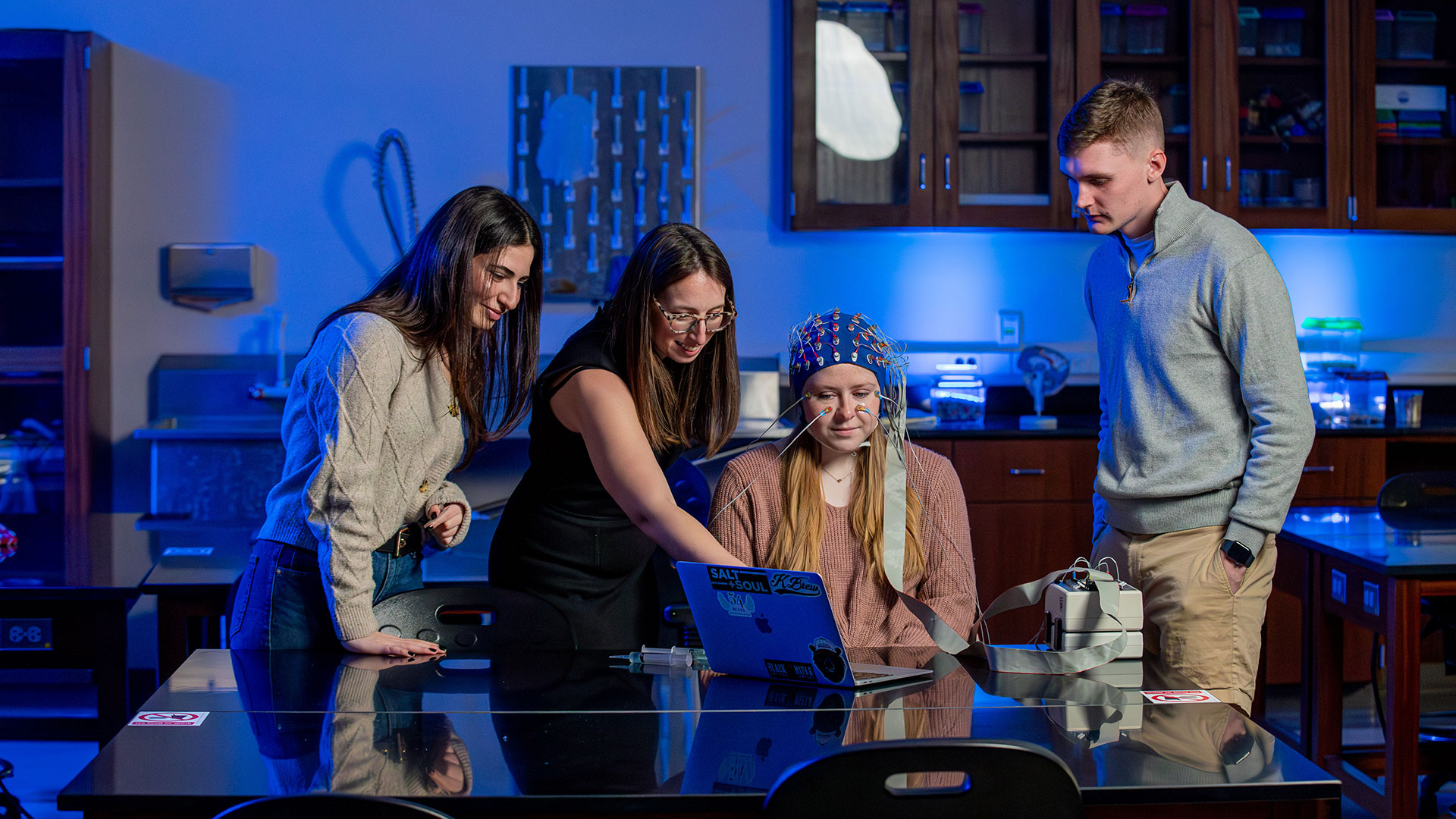 A female student wears a cap for an EEG while a professor explains the results to two other students