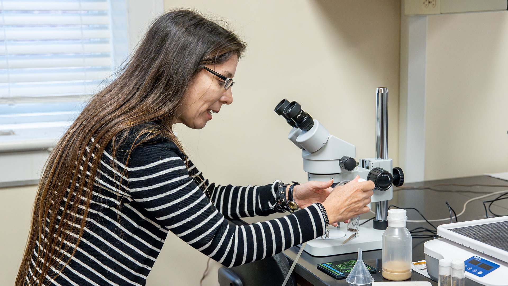  A woman with long hair and a striped shirt works with a microscope and lab equipment in a brightly lit science laboratory.