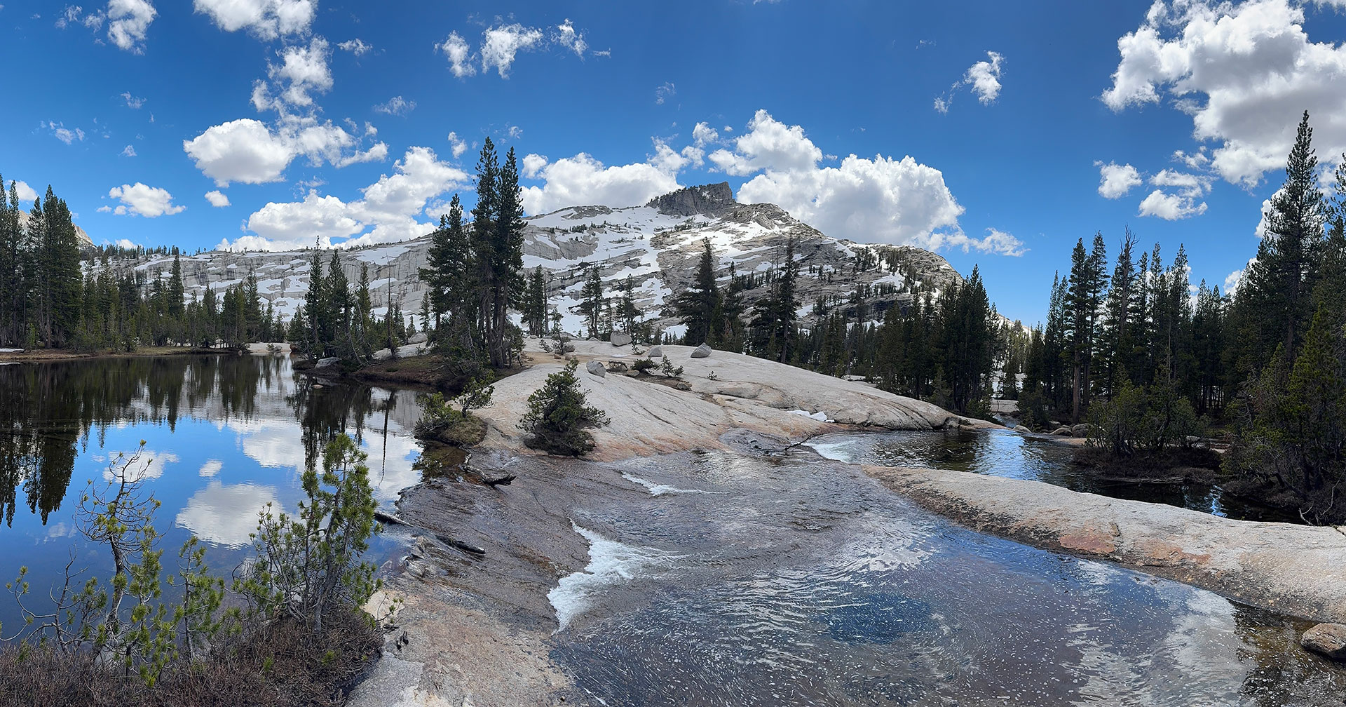 A sparsely snow-covered mountain with pine trees growing sparingly. In the foreground is water