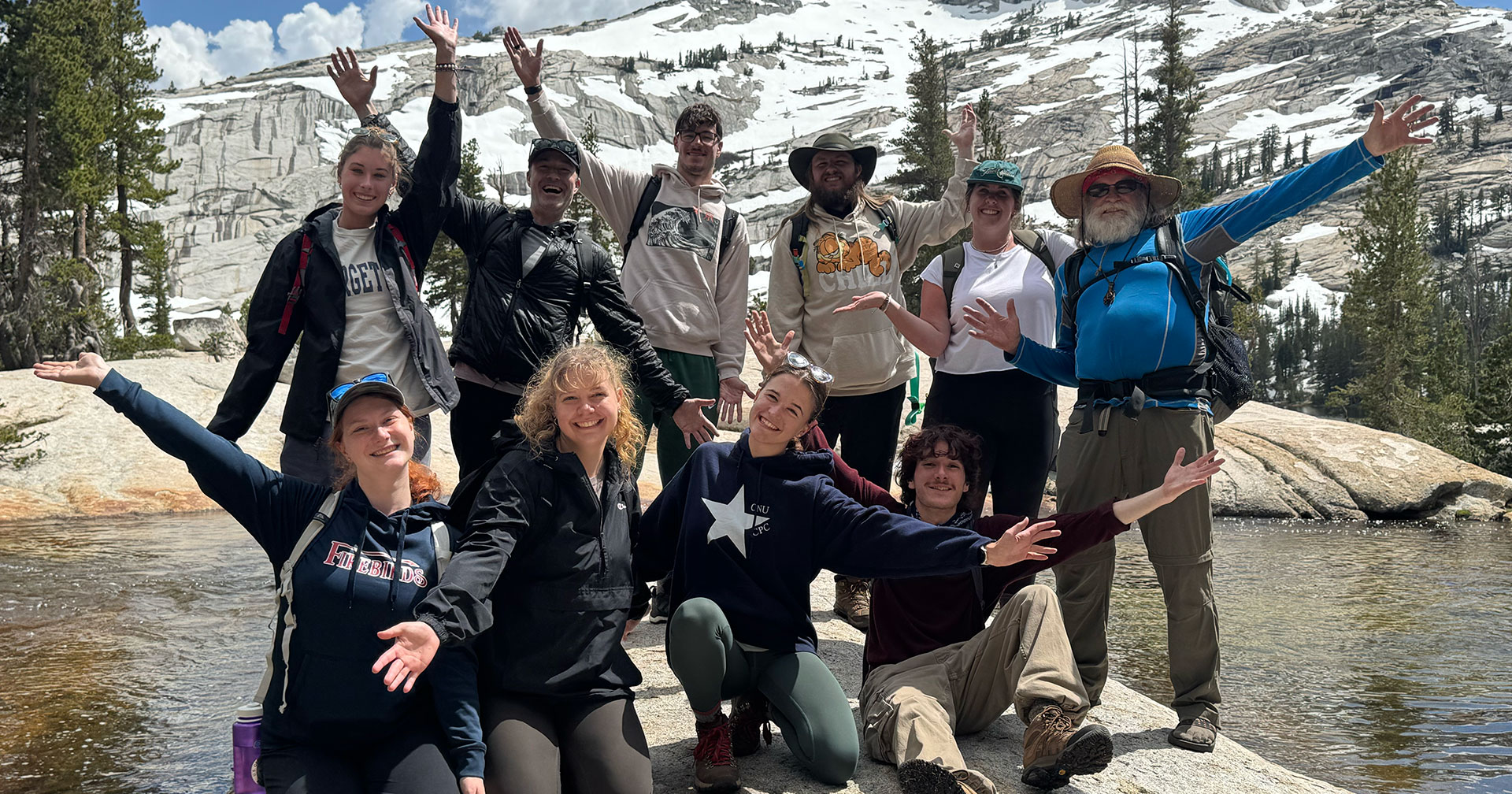 A group of people stand with arms outstretched amongst water and snow capped mountains in the background.
