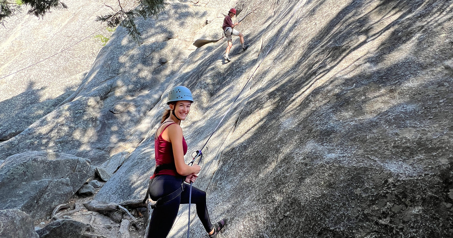 A woman wearing hiking gear smiles while standing semi-vertically on rock