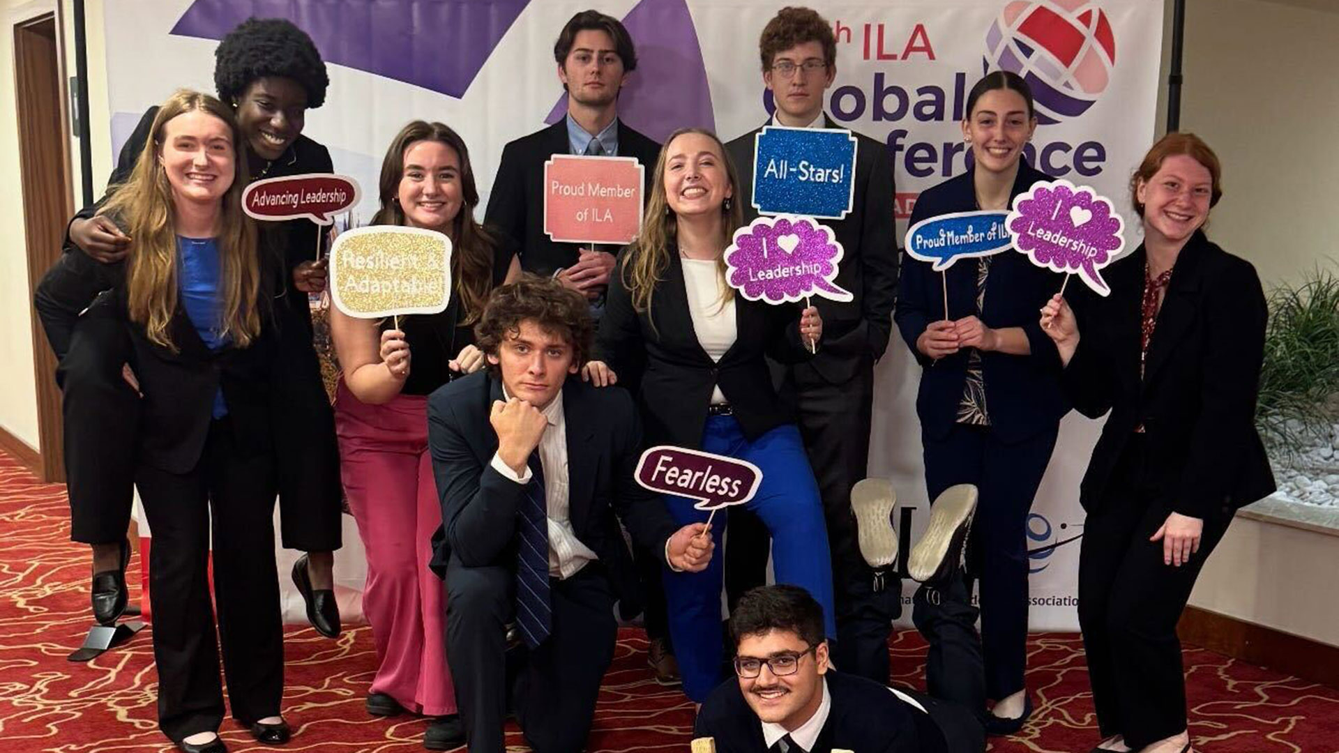 A group of nine diverse students, mostly young adults, dressed in business attire (suits and blazers), posing together indoors at an ILA Global Conference. They are holding up various glittery speech bubble signs that read: Advancing Leadership, Resilient and Adaptable, Proud Member of ILA, I heart Leadership, All-Stars!, Proud Member of ILA, Leadership, and Fearless. A tenth student is kneeling in front.
