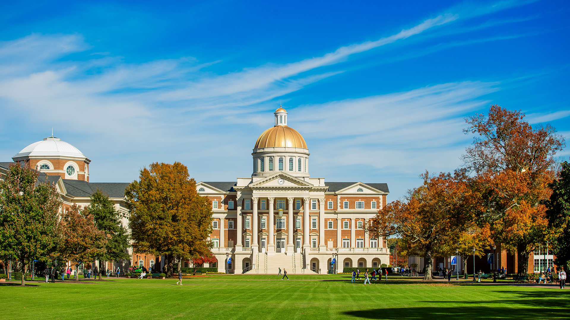 Christopher Newport Hall, a grand, classic university building with a gold dome and white columns, surrounded by a green lawn and trees with autumn foliage under a bright blue sky.
