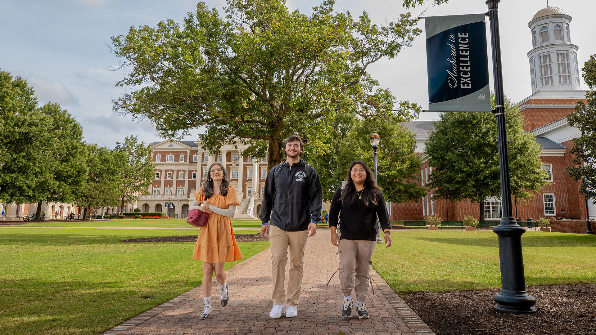 Three transfer students walk beside the Great Lawn next to a banner that reads Anchored in Excellence 