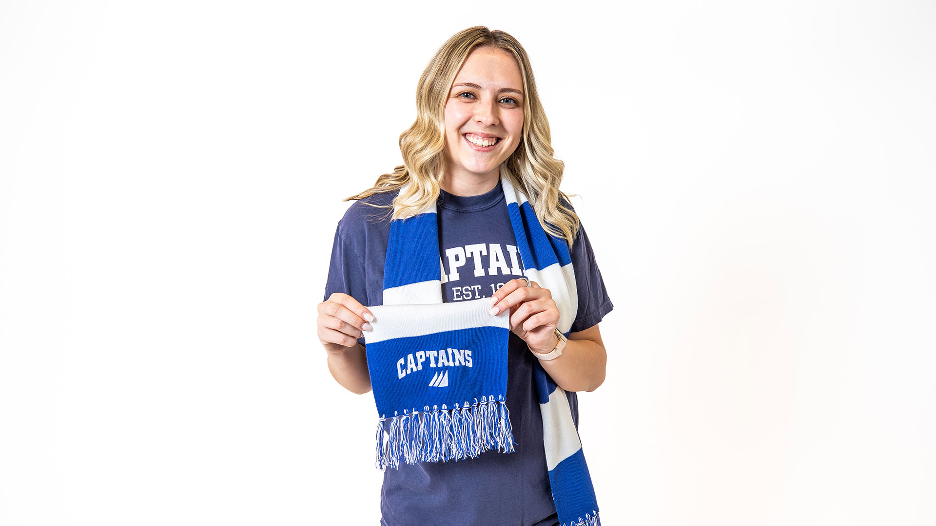 A student wears a blue and white striped scarf around their neck and is holding the end where the word Captains and the CNU 3 sails logo is embroidered.