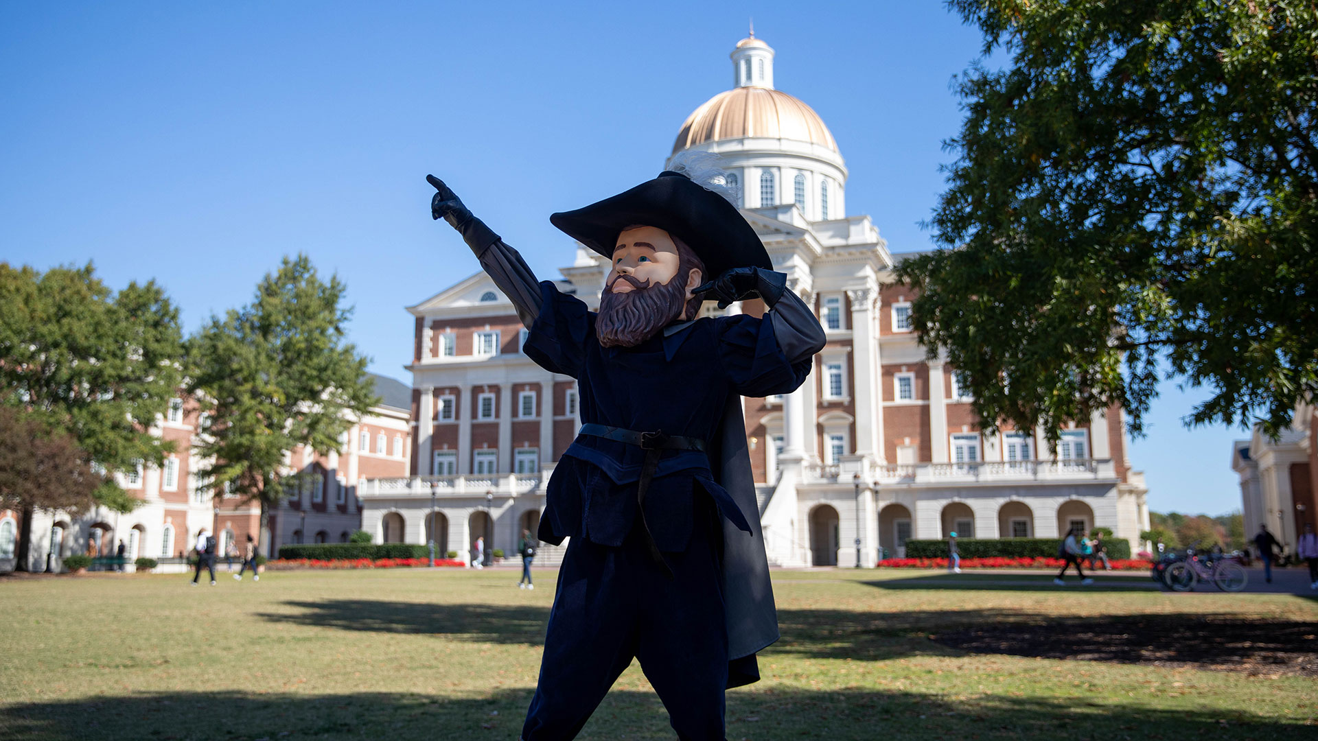 CNU mascot Captain Chris in a colonial outfit points toward a large domed building on a sunny university campus.