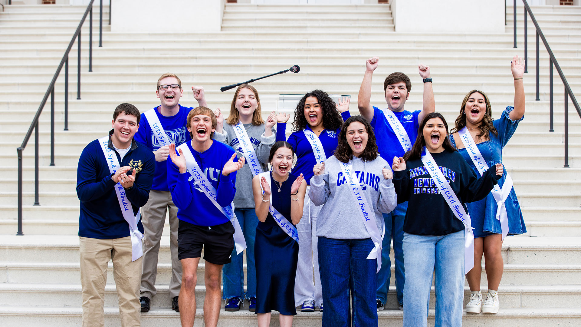 Students wearing white sashes with Spirit of CNU finalists in blue across their chests while wearing CNU spirit wear on the steps of Christopher Newport Hall